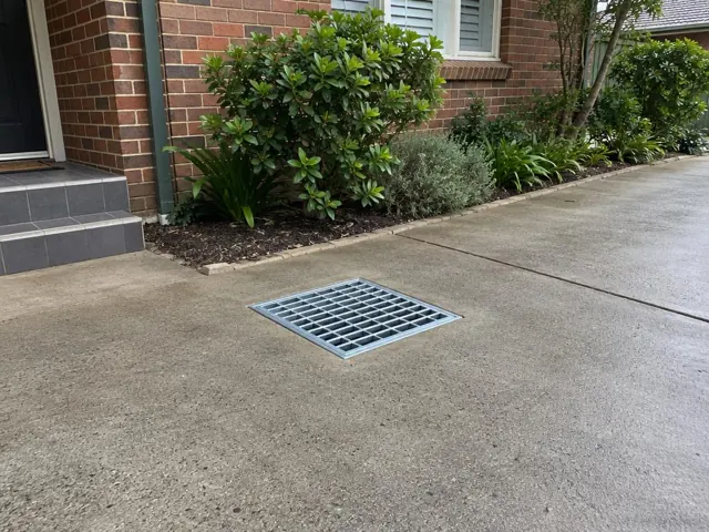 Flush stormwater grate in a concrete driveway beside a brick house in Waitara